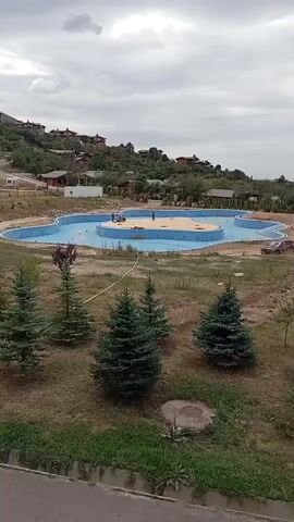 The video shows an outdoor scene with a large swimming pool under construction in the center of the frame. The pool is partially filled with blue water and has an unusual shape. There are some people working on the pool, and construction materials are visible nearby. The pool area is surrounded by a grassy landscape with several evergreen trees in the foreground. In the background, there are buildings situated on a hillside, and a parking lot filled with cars. The sky is cloudy. The camera appears to be panning slowly from left to right, capturing the entire scene.