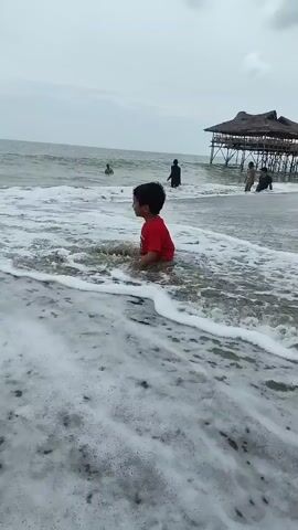 A young child wearing a red shirt sits on a sandy beach as gentle waves wash over them. The water creates foam and ripples around the child's legs. In the background, a stilted wooden structure with a thatched roof stands over the ocean. Several other people are visible in the water near the structure, some appearing to be children or younger individuals. The sky is overcast, suggesting a cloudy day. The overall scene depicts a relaxed beach environment with a focus on the child's interaction with the water.