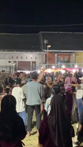 A group of people are gathered outdoors at night, likely at an event or festival. The background shows buildings with lights and decorations. In the center of the frame, a person is dancing in a sparkly outfit, raising their arms in the air. Many people are watching and some are reaching up towards the dancer. The ground appears to be grass.