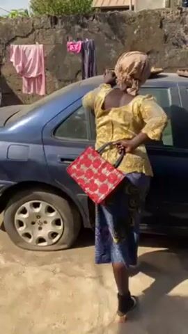 A woman stands beside a blue car with a flat tire. She wears a yellow blouse, a blue patterned skirt, black shoes, and a head wrap. She holds a red purse. The car has two trays of food on its roof. Laundry hangs on a line in the background near a stone wall.