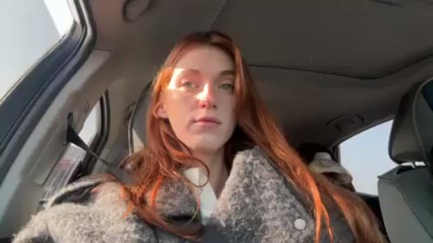 A woman with long auburn hair sits in the driver's seat of a car. She is wearing a gray wool-like jacket and a white shirt. The sun shines on her face. The car has a sunroof. A small person, likely a child, sits in the backseat.