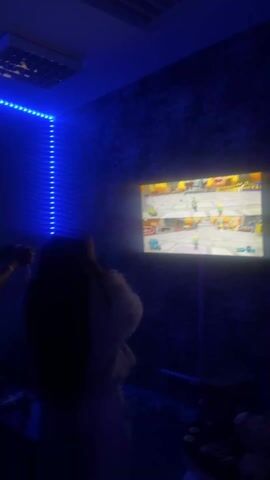 In a dimly lit room illuminated by blue LED strip lights, three individuals are engaged in a game of beer pong. The game is set up on a ping pong table with red plastic cups arranged at each end. The room's walls are painted in a dark hue, adding to the subdued ambiance. One of the individuals, a man, is dressed in a white t-shirt. The other two figures appear as dark silhouettes due to the low lighting. A glass bottle is visible on the edge of the table.