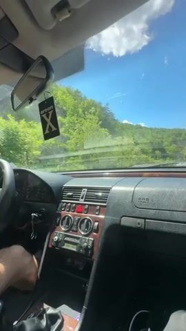 The video shows a view from inside a car driving on a highway. The dashboard is visible and features a wood grain trim. The road is surrounded by trees and green foliage, and the sky is clear and blue with some white clouds. As the car moves, the view shifts to the side window, revealing the roadside with trees and a metal guardrail. The car continues driving along the highway. The music appears to be dance-pop and is fairly loud.