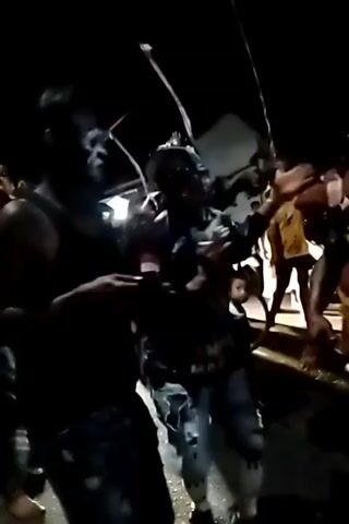 The video shows a group of people in a dark, outdoor setting. Some individuals are wearing face paint that resembles a skull or animal mask. The group appears to be engaged in some form of celebration or performance, with some individuals dancing or moving rhythmically. There are structures in the background that suggest an outdoor event or gathering. The lighting is dim, with some artificial lights illuminating parts of the scene.