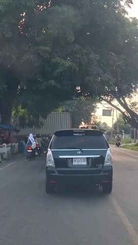 The video shows a black Toyota Innova driving on a road. The car has a license plate that reads BK 1445 GB. There are several motorcycles and people on the side of the road. A large tree is on the left side of the screen, and the sun is setting in the background.