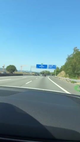 The video shows a first-person view from inside a car driving on a highway under a clear blue sky. The camera is positioned on the dashboard, facing forward through the windshield. In the foreground, the textured black dashboard of the car is visible. The road ahead is a multi-lane highway with white lane markings. Several cars are visible in front, driving in the same direction. On the right side of the highway, a metal guardrail runs along the edge, and beyond that, a grassy embankment covered with trees. On the left side of the highway, there are trees and some distant hills. Prominently, there are large blue highway signs overhead. One sign indicates "E-15 AP-7 Barcelona" with downward arrows. Another sign to the right indicates "500 m Cardedeu La Roca" and other smaller text. A construction crane is also visible in the far distance on the left. The overall environment appears to be a sunny day in a semi-rural or suburban area.