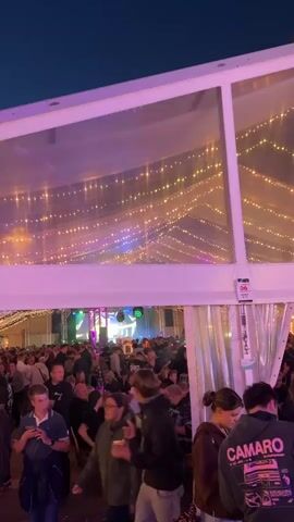 The video shows a crowded outdoor event at night. In the foreground, a group of people are gathered under a white tent with a clear roof covered in string lights. The tent has purple lighting on its sides. Inside the tent, a DJ booth with green and blue lights is visible, and many people are standing and socializing. To the right of the tent, people are standing around the entrance. In the background, there is a tree and further away, carnival rides with flashing lights are visible. The sky is dark blue.
