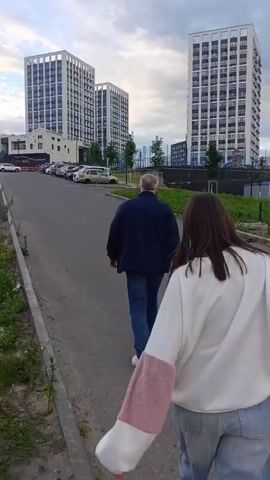 TWO PEOPLE, A MAN AND A WOMAN, ARE WALKING AWAY FROM THE CAMERA ON A PAVED PATH. THE MAN IS WEARING A DARK JACKET AND BLUE JEANS. THE WOMAN IS WEARING A WHITE SWEATSHIRT AND BLUE JEANS. THEY ARE WALKING TOWARDS THREE MODERN HIGH-RISE BUILDINGS THAT ARE PREDOMINANTLY WHITE WITH DARK WINDOWS. A FEW CARS ARE PARKED ON THE LEFT SIDE OF THE PATH. THE SKY IS CLOUDY. THE FOOTAGE APPEARS TO BE FROM A HANDHELD CAMERA, WITH THE CAMERA APPEARING TO BE SLIGHTLY JAGGED AS IT FOLLOWS THE SUBJECTS.