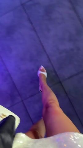 The video opens with a close-up of a person's legs and feet, wearing white heeled shoes and a sparkly dress. The camera then pans across a dimly lit room with blue and purple lighting. The scene shifts to reveal a party or event setting with several people seated at tables. The environment appears to be a modern indoor space, possibly a restaurant or event venue, with contemporary furniture and lighting fixtures. The overall atmosphere suggests a social gathering or celebration.