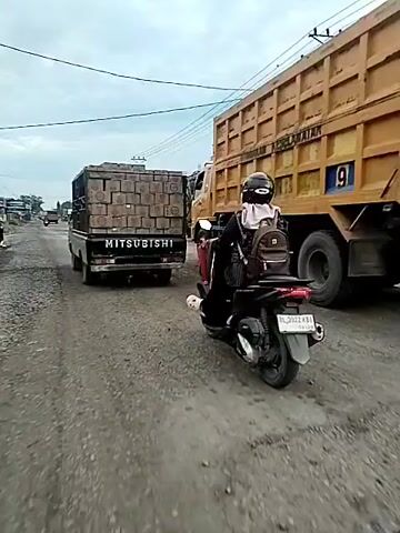 The video shows a dusty, unpaved road with several vehicles. In the foreground, a person on a scooter with a backpack is riding alongside a black Mitsubishi pickup truck loaded with what appear to be bricks or blocks. Behind the pickup truck is a large yellow dump truck. The road is lined with gravel and there are some buildings and power lines visible in the background. The sky is overcast. The scooter is in motion and appears to be passing the pickup truck. Another person on a scooter is visible further down the road to the right. The overall impression is of traffic on a rural or developing road.