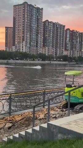 A person is water skiing on a river. The person is in the center of the frame, leaning back and creating a wake of white water behind them. They are wearing dark shorts and a dark top. In the background, there are tall buildings with many windows, suggesting an urban setting. Trees line the riverbank. The water is a dark, murky color. In the foreground, there is a metal railing and a green piece of machinery that appears to be a small excavator. The sky in the background is a soft pink and orange, indicating that it is either sunrise or sunset. The overall mood is one of recreation and leisure. There is no visible text in the video.
