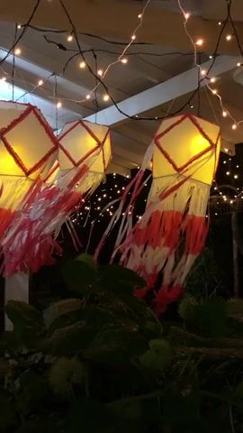 The video shows a low-angle view of a collection of Vesak lanterns hanging under a white ceiling, adorned with string lights. The lanterns are diamond-shaped with red and white fringe hanging from the bottom, and they glow with a warm, yellow light. The background is dark, punctuated by more string lights that appear as small, blurry dots. The bottom of the frame includes dark green foliage. The camera remains static throughout the video, providing a still, celebratory scene.