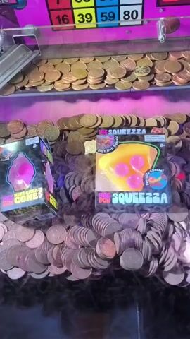This is a close-up, vertical shot of a coin pusher arcade game, likely in an amusement center. In the foreground, a large pile of golden and copper-colored coins fills the bottom of the screen, some of them in motion. Two prizes, one a pink ice cream cone-shaped object in a box labeled "MEGA ICE CREAM CONES," and another a yellow pizza-shaped object in a box labeled "MEGA SQUEEZZA," are positioned near the center of the coin pile. Behind the coins and prizes, a mechanism with a metal chute is visible at the top, from which more coins appear to be falling. Above this, a bright pink panel with a grid of colored squares containing numbers is displayed. The squares are arranged in rows and columns, and some have red or blue numbers. A digital red display above the number grid shows the number "88." The lighting is bright and colorful, with a mix of purples and yellows illuminating the scene. The overall impression is that of an active arcade game in progress.