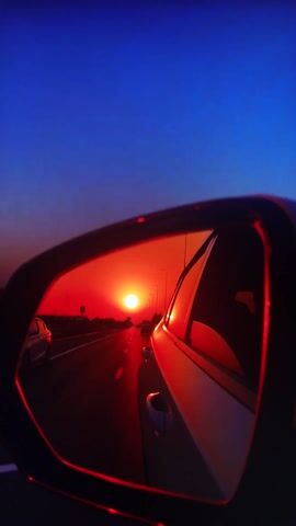 This media file is a close-up, slow-motion shot of a wing mirror of a vehicle, likely a car or truck, at what appears to be sunset or sunrise. The mirror reflects a road stretching into the distance, lined with trees. The sun is a bright, reddish-orange orb, casting a warm glow on the scene. A few vehicles are visible on the road, also reflecting the sunset. The sky in the background is a deep, twilight blue, creating a stark contrast with the fiery hues in the mirror. The overall mood is serene and contemplative, with a sense of movement and passage suggested by the reflection of the road. There is no visible text in the media file. The location context provided places this scene in Belorechensk, Russia.