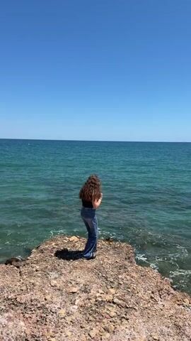 The video shows a young person with long, dark curly hair, wearing a black tank top and blue bell-bottom jeans, standing on a rocky outcrop by the sea. The person is facing the water and moves their arms as if dancing. The sea is a clear turquoise color and the sky is bright blue. The person smiles and laughs at the camera. The video is shot from a slightly low angle.