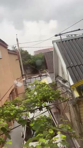 A bird's eye view of a residential area under a cloudy sky. Buildings with tiled roofs and light-colored walls are visible, some with air conditioning units mounted on them. A satellite dish is attached to one of the walls. Green foliage from trees and vines is interspersed between the structures. Power lines and a utility pole run through the scene. The overall impression is one of an overcast day.
