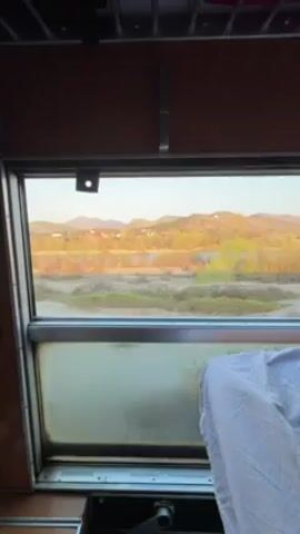 The video shows a view from inside a train car looking out of a window. The window is slightly fogged and has metal framing. Outside the window, the landscape is visible in the late afternoon or early evening light. The landscape features scrubby vegetation, a river, and some buildings reflecting the warm light. In the distance are hills or mountains. Inside the train car, part of a bed or seat with a white cloth covering is visible.