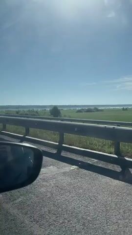 The video shows a view from inside a car driving on a road, likely a bridge, under a bright, sunny sky. The road is gray asphalt.  A gray guardrail lines the right side of the road.  Beyond the guardrail is green grassy vegetation and further beyond that, what appears to be a body of water with some land visible in the distance. The side mirror of the car is visible in the lower left corner of the frame.