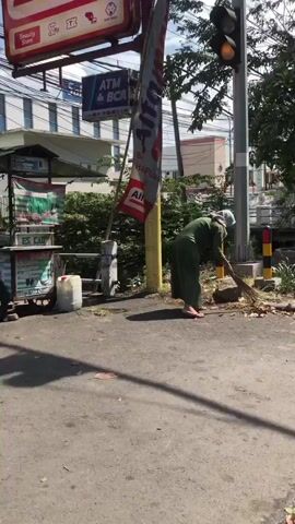 A woman in a green hijab and matching green outfit is sweeping leaves and debris from an asphalt road with a broom. She is standing on the right side of the frame, facing left, and is bent over the pile of leaves. The background shows a building with signs, a lamppost, and lush green foliage. The scene appears to be outdoors during the day.