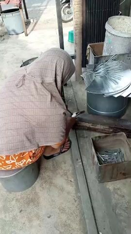 A person wearing a headscarf and an orange patterned skirt is sitting on a large silver cylindrical object, facing away from the camera. The person is bent over a workbench, and appears to be working with metal. There are various metal objects and tools scattered around the person, including a hammer, a pile of metal sheets, a can of wire, and a metal box filled with small metal pieces. The scene takes place outdoors, on a concrete surface, with a metal fence or gate in the background.