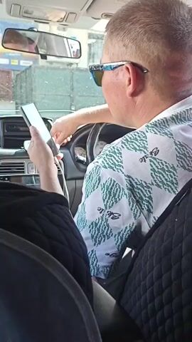 The media file shows the interior of a car from the perspective of a passenger. The main subject is the driver, a man wearing sunglasses and a white shirt with a green diamond pattern. He is looking at his phone, which he holds in his left hand. The car's interior is visible, including the dashboard, steering wheel, and rearview mirror. The scene appears to be taking place during the daytime, with natural light entering the car. The outside environment is blurred but suggests a somewhat overcast or rainy day, with water droplets visible on the window. The location is identified as Kazarman, Kyrgyzstan. No specific emotions are clearly discernible from the subjects, but the act of looking at a phone while driving suggests a potential distraction. There is no visible text in the media file.