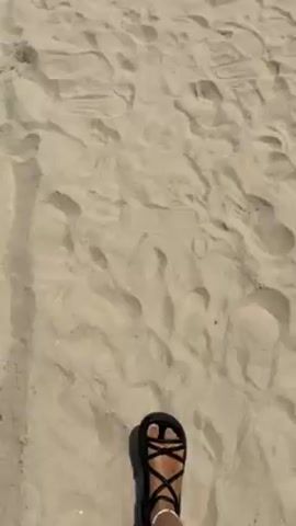 The video shows a sandy beach. The camera pans up from the sand to show the ocean and sky. The ocean is greenish-blue and has small waves. The sky is a clear blue with a slight haze. There are many footprints in the sand.