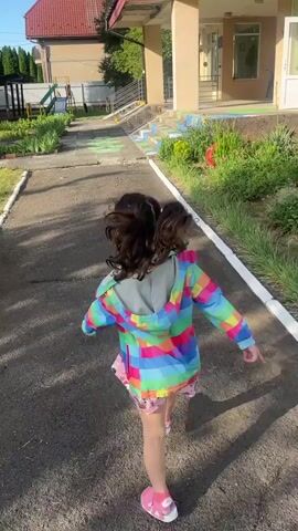 A young girl with dark hair in a ponytail runs down a paved path in front of a building. She is wearing a colorful jacket and a patterned skirt. The path is bordered by grass and white lines. In the background, there is a building with a red roof and a playground with a slide. The sun is shining.