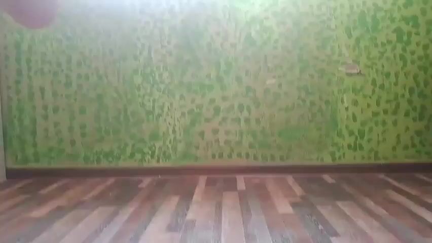 The video shows a sparsely furnished room with a distinctive green and tan textured wall and a dark wood floor. The camera pans across the wall, revealing peeling paint in some areas. The view then shifts to an outdoor scene, looking out between two apartment buildings. The courtyard below contains a white car, some trees, and other buildings in the distance.