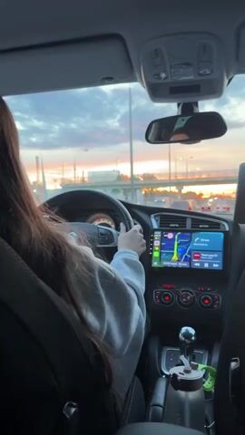Here is a description of the video:

The video shows a woman driving a car. She has long brown hair and is wearing a gray sweatshirt. The car has a digital display screen showing a map and navigation information. The sky outside is visible through the windshield and shows a sunset with orange and blue hues. There is a bridge in the distance. The interior of the car is mostly black. The video is shot from the backseat.