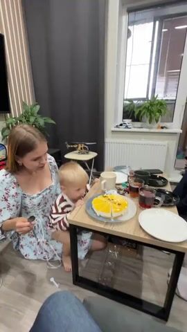 A young woman in a floral dress holds a blonde toddler on her lap at a small wooden table. A yellow cake with a passion fruit topping is on a plate in the center of the table. The woman is holding a fork and appears to be cutting or serving a piece of the cake to the toddler, who is looking at the cake. Two mugs of dark liquid, presumably tea, are also on the table, along with plates and other items. The scene appears to be indoors, with a window and plants visible in the background. The toddler is wearing a striped shirt.
