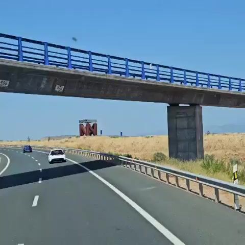 A wide-angle shot captures a highway scene under a clear blue sky. On the right, a highway guardrail runs alongside the road. To the left, a white car travels ahead, with another vehicle, possibly blue, further in the distance. A concrete overpass with blue railings spans the highway. In the background, a dry, grassy field stretches out. Prominently featured in the middle ground is a large, abstract sculpture made of rusty metal, resembling two large "N" shapes supporting a flat, metallic top. Two blue signs are visible behind the sculpture. The overall environment suggests a sunny, arid landscape.