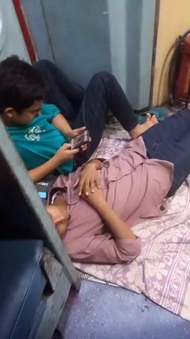 The video shows four people lying on the floor of a train car. Two are lying on a blanket with a purple floral pattern, and two are lying on a brown blanket. The train car has a metal floor with a diamond pattern, blue walls, and yellow accents. There are various items in the train car including a fire extinguisher, a trash can, and luggage.