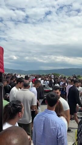 The video shows a large crowd of people gathered on an open road, likely for an event of some kind. Most of the people are young adults, both male and female, dressed casually in t-shirts, shorts, jeans, and various styles of tops. There's a diverse mix of colors in their clothing, with black, white, and light colors being prevalent.

In the foreground are several individuals who appear to be observing the activity ahead. The focus is on the crowd stretching into the distance towards a horizon of rolling hills and mountains under a partly cloudy sky. A few cars are visible among the crowd. 

The overall atmosphere appears to be lively and festive. The event seems to be taking place outdoors in a rural or semi-rural setting.