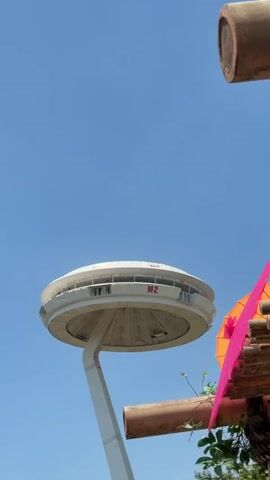 The video is a vertical shot of a futuristic-looking building on a clear, sunny day. The building is white and circular with windows around its perimeter. It is supported by a tall, angled white pillar. The words "M2" are visible in red on the side of the building. In the foreground, there are some wooden structures and greenery, as well as a sign with Italian text. Several people are walking in the background on a paved pathway. The sky is a bright blue.