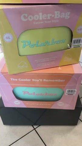 The video shows a stack of pink and yellow boxes for Polarbox cooler bags. The boxes have text in English and French, and depict images of the cooler bags. The background is blurry but appears to be a retail store with shelves of merchandise. A price list for the Polarbox cooler bags in euros is visible on a stand to the right of the boxes. The cooler bags themselves are pink and have "Polarbox" written in yellow lettering on them. The video pans up and down the stack of boxes, showing different sizes and colors of the cooler bags. The lighting is bright and appears to be artificial.