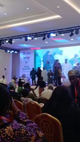 This video captures an indoor event, likely a conference or seminar, in a large hall. On a brightly lit stage, a group of approximately ten individuals, dressed in formal and traditional attire, are standing together, possibly posing for a photograph or after a presentation. Behind them, a large screen displays the event's branding, which includes "NRRW Croc City 2023" and the theme "Food Security". Below the main title, "Lale-La" is also visible. The screen also shows what appears to be a larger version of the people currently on stage, suggesting a previous visual or a backdrop.

In the foreground and middle ground, an audience is seated in rows of chairs, mostly facing the stage. Many attendees are wearing traditional Nigerian clothing. Some audience members are looking towards the stage, while others are looking down or engaged with their phones. The overall atmosphere appears formal and attentive. The venue is equipped with stage lighting and a sound system, indicated by the spotlights and speakers mounted above the stage. The event seems to be focused on food security, taking place in "Croc City" (likely referring to Kaduna, Nigeria, consistent with Unguwan Sanusi's location) in the year 2023.