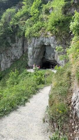 The video starts with a close-up shot of a cave entrance set into a rocky cliff face, surrounded by lush green vegetation. A small group of people is gathered near the cave entrance. The camera then pans upwards and outwards, revealing a broader landscape of rolling green hills, forested areas, and distant mountains under a sky with some scattered clouds. Power lines stretch across the scene. The overall impression is one of a mountainous, natural environment.