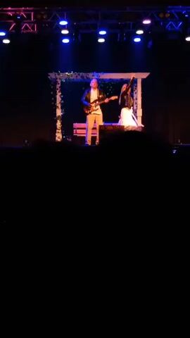 Here is a detailed description of the scene:

The video depicts a live music performance taking place on a stage. A male musician is playing an electric guitar on the left, while a female singer stands to his right, singing into a microphone. The musician is wearing a black leather jacket over a white shirt, gray pants, and brown shoes. The singer is dressed in a white dress, black jacket, and blonde hair.

Behind the performers is a decorative white wooden frame covered in green vines, with a wooden bench placed in front of it. Above the stage, there are stage lights arranged on a metal structure, shining in various colors, including blue, white, and pink.

The scene is filmed from the audience's perspective, with silhouettes of people's heads visible in the foreground, indicating a crowd watching the performance. The lighting is dim, focusing on the stage and the performers, which suggests an indoor venue. The overall setting is a public event, likely a concert or show.