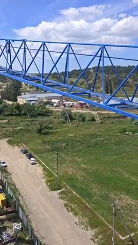 The video shows a view from a construction crane, looking down on a site under construction. The crane's blue boom arm extends across the frame. Below, there is a dirt road with several cars parked alongside. Construction materials and portable buildings are visible on the ground. In the background, there are trees and buildings, suggesting a semi-rural or suburban setting. The sky is blue with some white clouds. The crane is actively being assembled or disassembled, with parts being lifted and positioned.