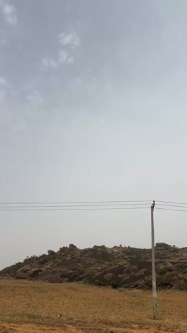The video shows a landscape under a sky partially covered with thin clouds. The ground is mostly dry, arid land with sparse vegetation. A rocky hill with larger boulders and some greenery is visible in the background. Power lines are stretched across the view, supported by utility poles. The overall impression is of a rural, possibly remote, area with a dusty or hazy atmosphere.
