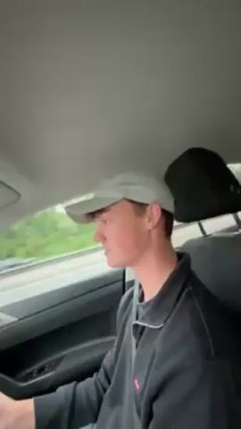 Here is a concise description of the video:

The video is shot from the perspective of someone in the driver's seat of a car, driving on a highway in wet conditions.  The driver is a young man wearing a light-colored baseball cap and a black sweater. The interior of the car is modern, with a digital display on the dashboard. Outside, rain streaks the windshield and other cars are visible, including a double-decker bus and a car carrier truck.