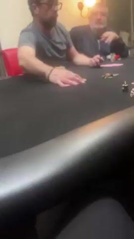 Two men are playing poker at a black poker table in an indoor room. The man on the left wears a grey t-shirt and glasses, and is pushing chips onto the table. The other man wears a blue jacket and has a beard. There are a few poker chips scattered on the table. A red chair sits behind the man on the left, and a lamp is on the wall behind the man on the right. The bottom of the video shows a black leather edge of the poker table.