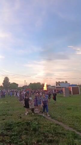 A large group of people are gathered outdoors, some walking along a dirt path in a grassy field. In the distance, a large bonfire is burning. The sky is cloudy with hues of orange and pink, indicating sunset. There are some trees, power lines, and buildings visible in the background. The people appear to be dressed in casual clothing, and the overall atmosphere seems festive. Some people are looking towards the bonfire, while others are walking or talking amongst themselves.