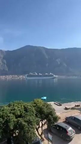 The video shows a large cruise ship on calm blue water with a mountain in the background. The ship is white with a blue and black logo, and "PRINCESS CRUISES" is written on the side. The mountain appears to be rocky and covered in vegetation. In the foreground, there are trees, a small blue raft floating on the water, a parking lot with a few cars, and a section of a building. The sky is blue and clear. The camera zooms in on the cruise ship.