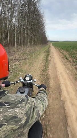The video shows a first-person view of a person riding a motorcycle on a dirt road. The rider wears a red helmet, a camouflage jacket, and black gloves. The motorcycle appears to be an older model with a black gas tank and chrome handlebars. The dirt road is flanked on one side by a row of tall, bare trees and on the other by a green field. The sky is overcast. The rider continues along the dirt road at a moderate pace.