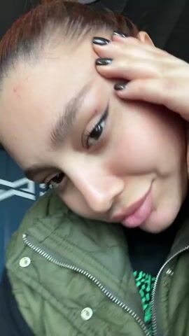 A close-up, vertically oriented shot shows a woman indoors. She is fair-skinned with light reddish-brown hair styled up. She's wearing an olive-green quilted vest over a black t-shirt with a green pattern. She is also wearing a grey smartwatch. She smiles slightly with her right hand on her forehead, and her fingernails are painted black. The background includes a dark blue paneled wall with a light switch, a white framed object, and a blurred ceiling light. The room appears to be well-lit.