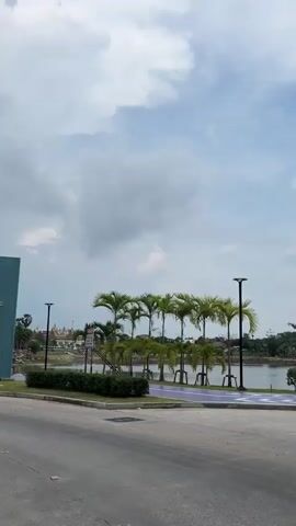 The video shows a sign in Thai for "Suan Sakon Nakhon", Phase 3. Behind the sign is a lake with palm trees planted along the shore. In the distance, across the lake, there are buildings with traditional Thai architecture. The sky is cloudy with patches of blue. A gray guardrail runs along the road in front of the lake.