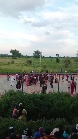 The video shows a group of people gathered on an outdoor court, possibly a tennis or basketball court, with a red and green surface. A fence separates the court from an area with dense green bushes in the foreground. Beyond the court, there is a grassy field with scattered trees and power lines. The sky is partly cloudy, and the lighting suggests it is either early morning or late afternoon. Many individuals on the court appear to be wearing traditional clothing, with some in vibrant colors. Some people are standing and observing, while others seem to be interacting or possibly dancing. A few individuals are visible in the foreground, partially obscured by the bushes, also appearing to watch the event.