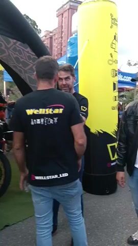 The video shows an outdoor event taking place on a cloudy day in what appears to be an urban setting. 

Several people are milling around a street area, with tents and promotional displays set up, suggesting a market or festival. There are individuals of varying ages and genders, dressed in casual attire. Some are seen talking to each other, while others are walking through the area.

Several motorcycles are also visible, one being maneuvered through the crowd. A yellow inflatable pillar with black branding is prominent in the foreground. There are also buildings in the background, adding to the urban backdrop.

The overall impression is a moderately crowded public event or gathering.