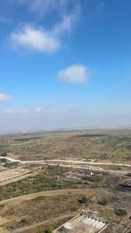 A drone flies over a vast, arid landscape under a bright blue sky with scattered white clouds. The terrain is a mix of dry grass, scrub brush, and patches of darker soil. In the mid-ground, a road winds through the landscape, with what appears to be an abandoned industrial site featuring concrete structures and rusting metal. Further in the distance, the landscape stretches to the horizon, with hints of vegetation and some buildings in the far distance. In the foreground, a rusty metal railing with yellow accents is visible, suggesting the drone is ascending from a structure. The overall impression is one of a desolate, undeveloped area with remnants of industrial activity.